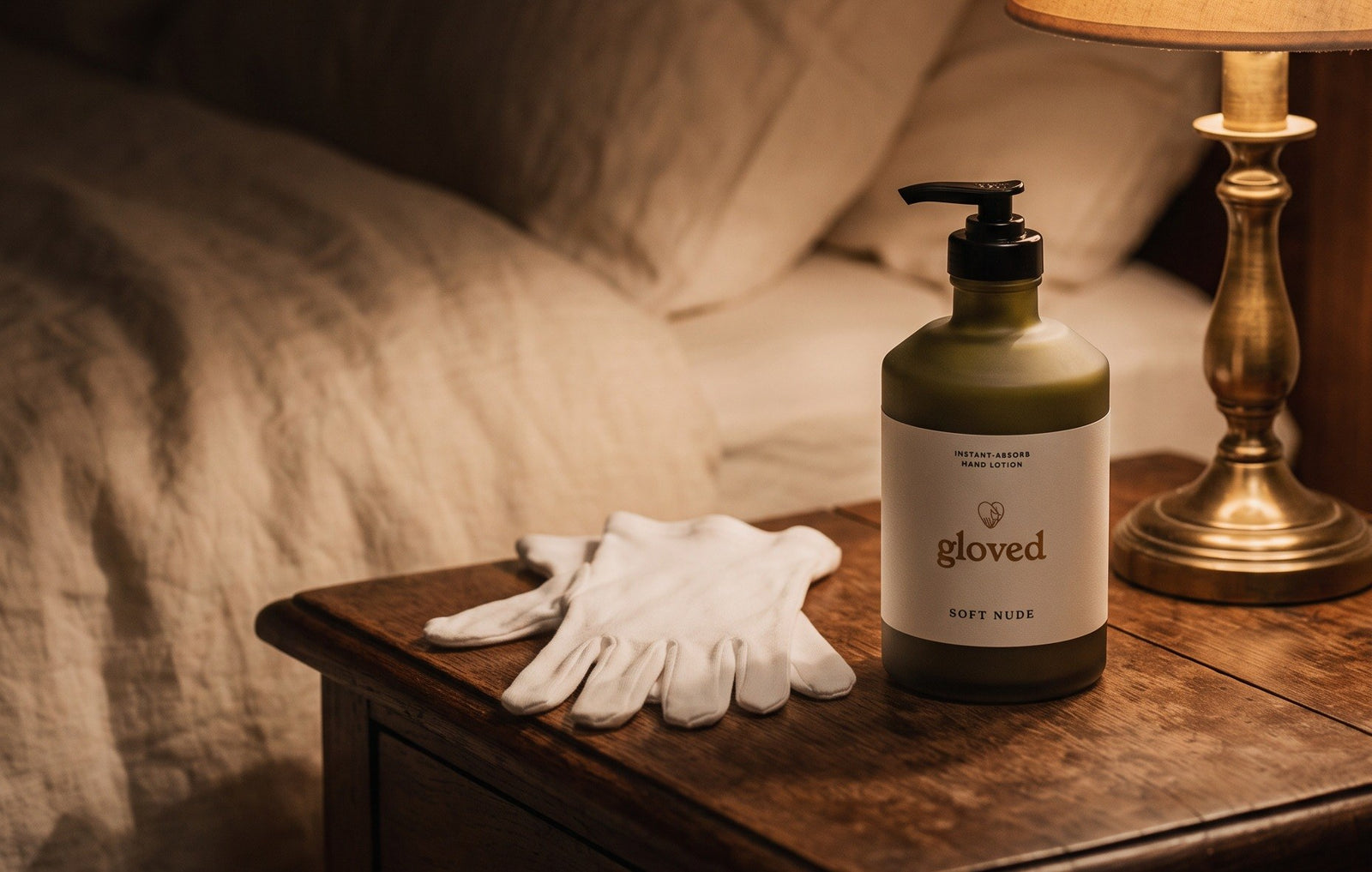 A bedside table at night: a lamp, a pair of folded white cotton gloves and the gloved Instant-Absorb Hand Lotion